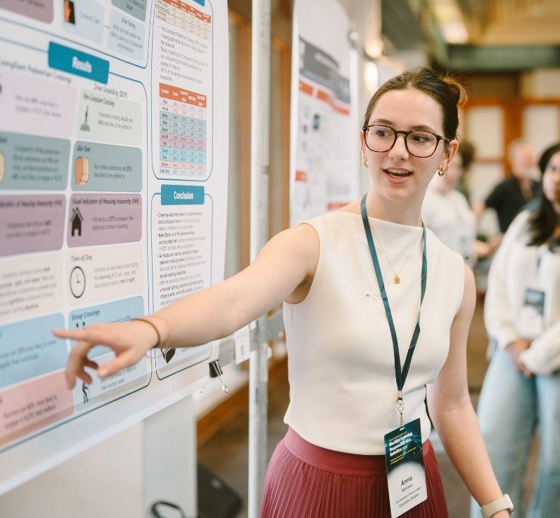 Student points to a research poster while explaining findings to other students.